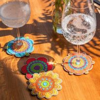 Sous-verres fleurs hippies Diamonds Dotz utilisés sous des verres sur une table en bois éclairée par la lumière naturelle. Les motifs mandala multicolores créent une ambiance chaleureuse, bohème et estivale.