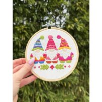 Broderie terminée tenue en main dans son tambour en bois de 15 cm. Le motif présente trois gnomes barbus portant de hauts bonnets colorés (bleu à motifs, rose et jaune/rose) placés au-dessus de feuilles de houx vertes et d'une petite fleur rose.