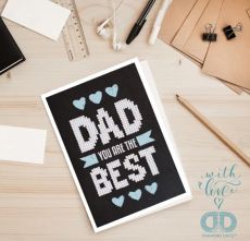 Vue de dessus d'un bureau en bois clair sur lequel repose la carte de vœux Diamond Dotz "Dad You Are The Best". On aperçoit autour de la carte des accessoires de papeterie : une règle, des écouteurs blancs, un crayon à papier et des pinces à dessin.